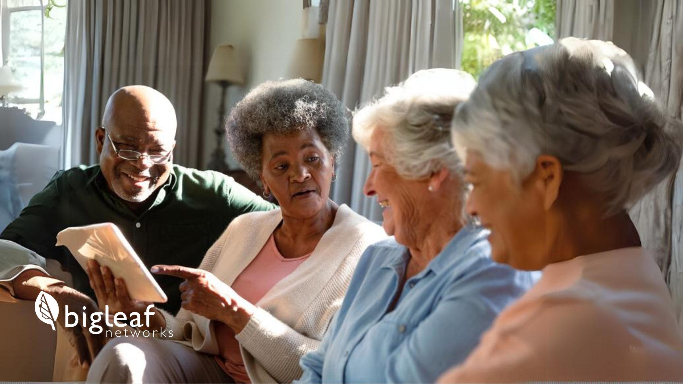 A group of four senior friends, including three women and one man, are joyfully interacting with a tablet while sitting on a sofa.