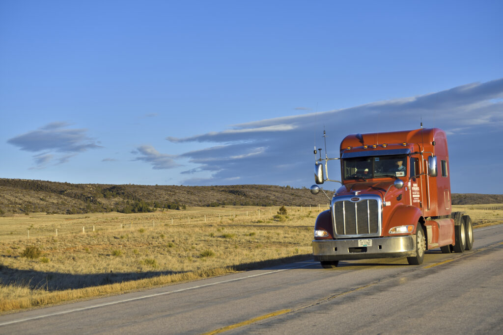 Red Peterbilt truck driving on a scenic highway, representing the fleet and operational reach of Allstate Peterbilt Group in the upper Midwest.