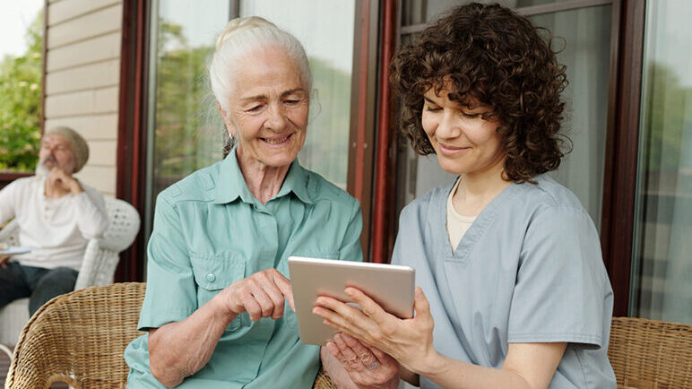 Elderly woman with caregiver learning to use a tablet on a sunny assisted living facility porch, with senior men conversing in the background.