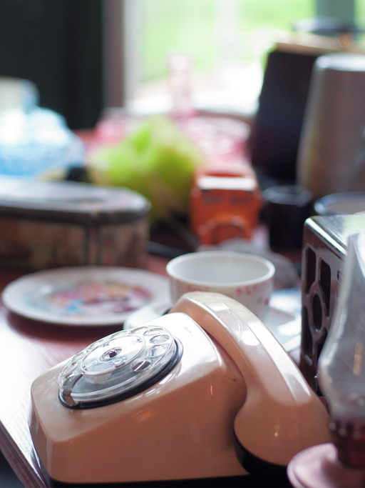 Close-up of a vintage rotary telephone amidst a softly blurred background of household items and a window that suggests a cozy, lived-in space.